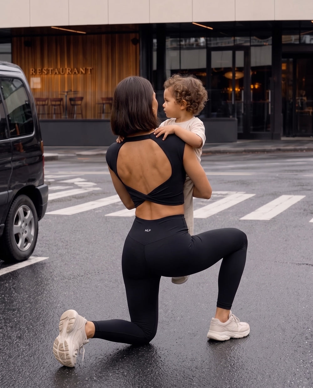Mom in the black Knot top carrying her toddler across the street at pickup